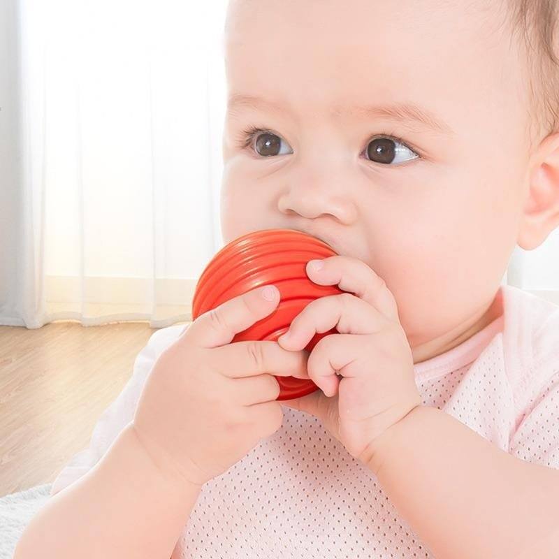 A baby with a thoughtful expression chewing on a red InvenToy Montessori Shape Blocks, holding it with both hands, in a brightly lit room with a sheer curtain in the background.