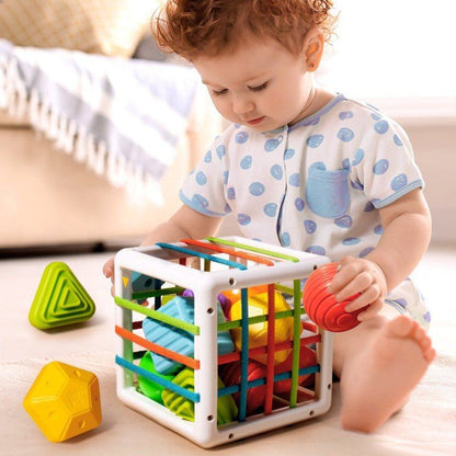 A toddler with curly hair, wearing a blue polka dot outfit, playing with a colorful InvenToy Montessori Shape Blocks toy on a sunny, carpeted floor.