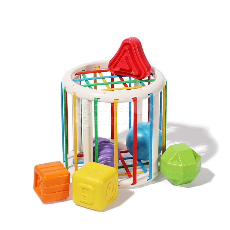 A colorful InvenToy Montessori Shape Blocks set featuring a white cage with multicolored bars and a red top, surrounded by geometrically shaped blocks in orange, yellow, purple, and green.