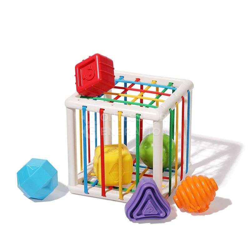 A colorful plastic playpen with multicolored vertical bars contains a red shape-sorter lid and various InvenToy Montessori Shape Blocks scattered around it, on a