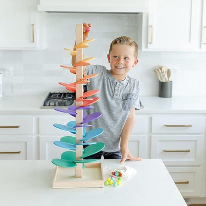 A young boy smiles at the camera while playing with a Montessori Rainbow Tree in a bright kitchen. He is about to drop a marble onto the spiraling track designed by InvenToy.